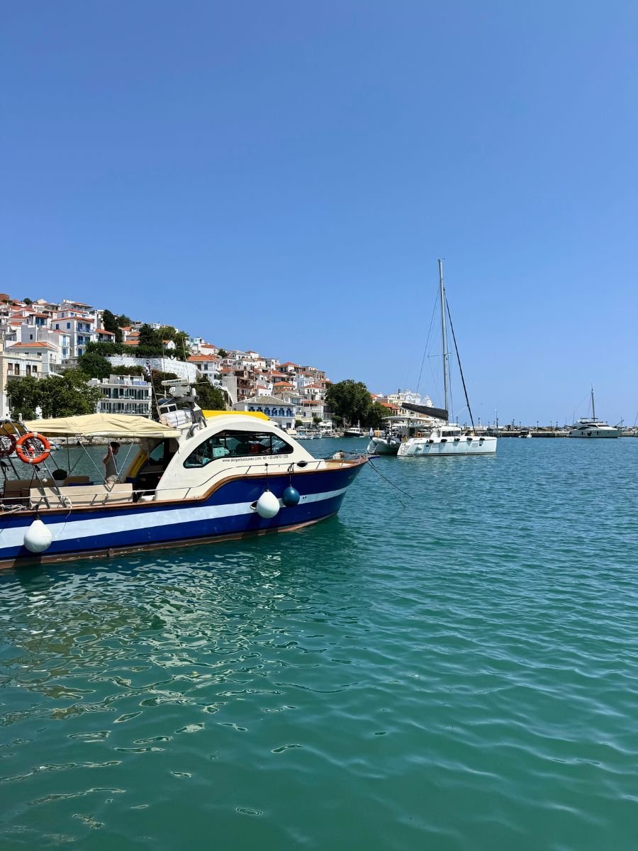 Colorful boats anchored in the turquoise waters of Skopelos harbor, with whitewashed hillside houses and a clear blue sky in the background.