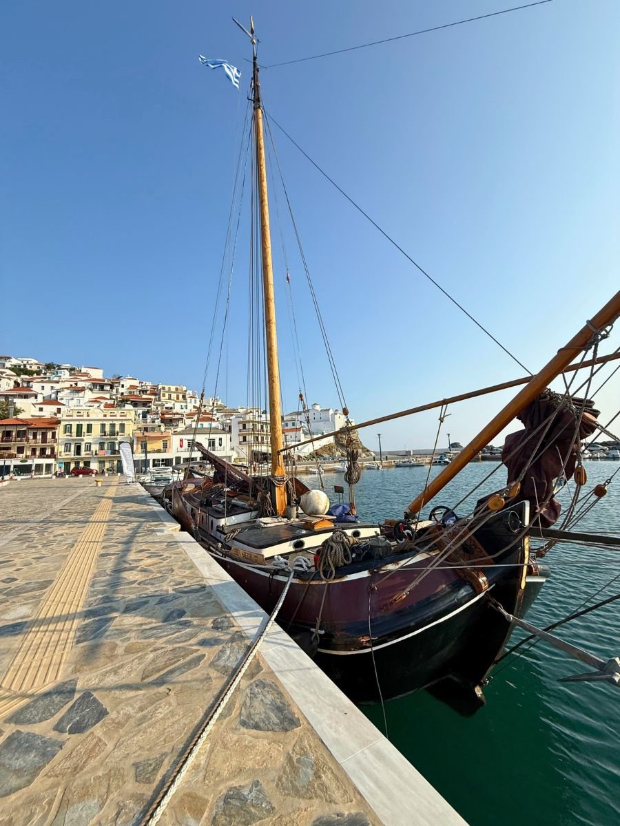 Traditional wooden sailboat docked at the harbor in Skopelos, Greece, with colorful hillside buildings and a clear blue sky in the background.
