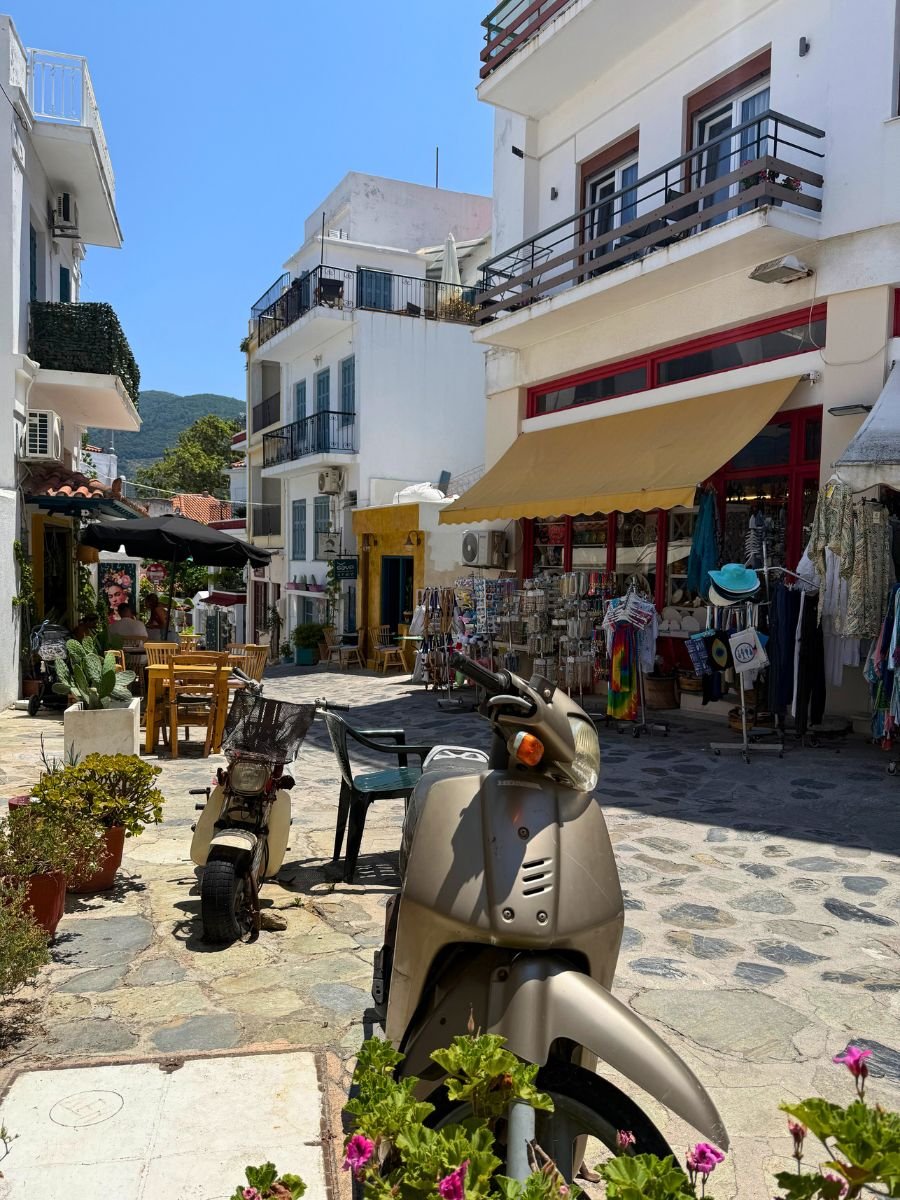 Charming street in Skopelos town with parked scooters, local shops, and outdoor café tables on a sunny day.