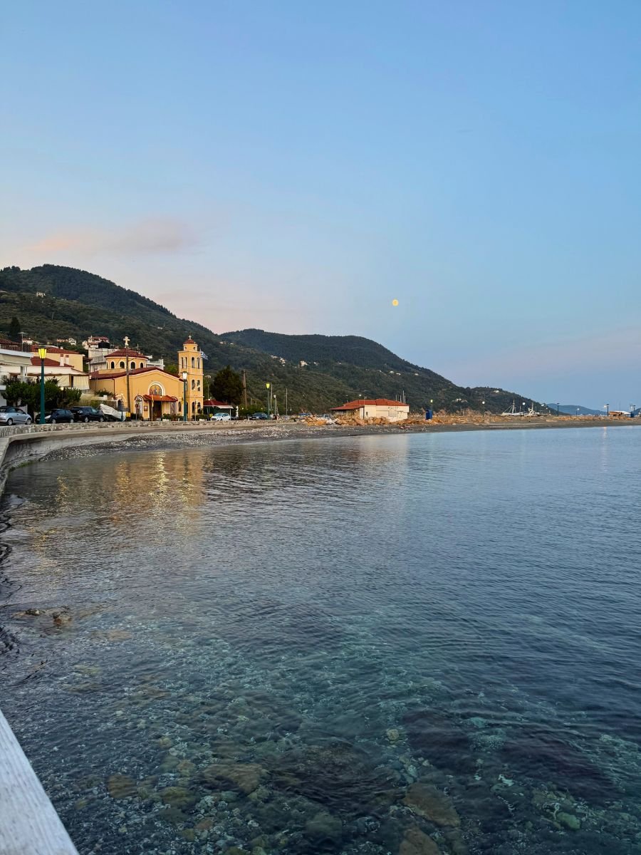 Peaceful coastal view of Skopelos Town at sunset, with clear blue water, distant mountains, and a yellow church by the shoreline.