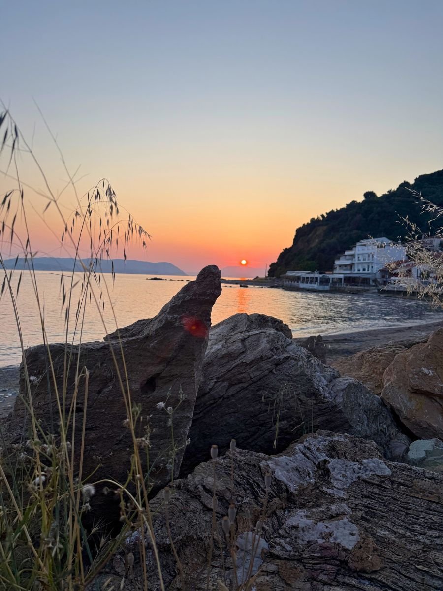 Sunset over the Aegean Sea in Skopelos, with rocky foreground and silhouettes of hills and seaside buildings.
