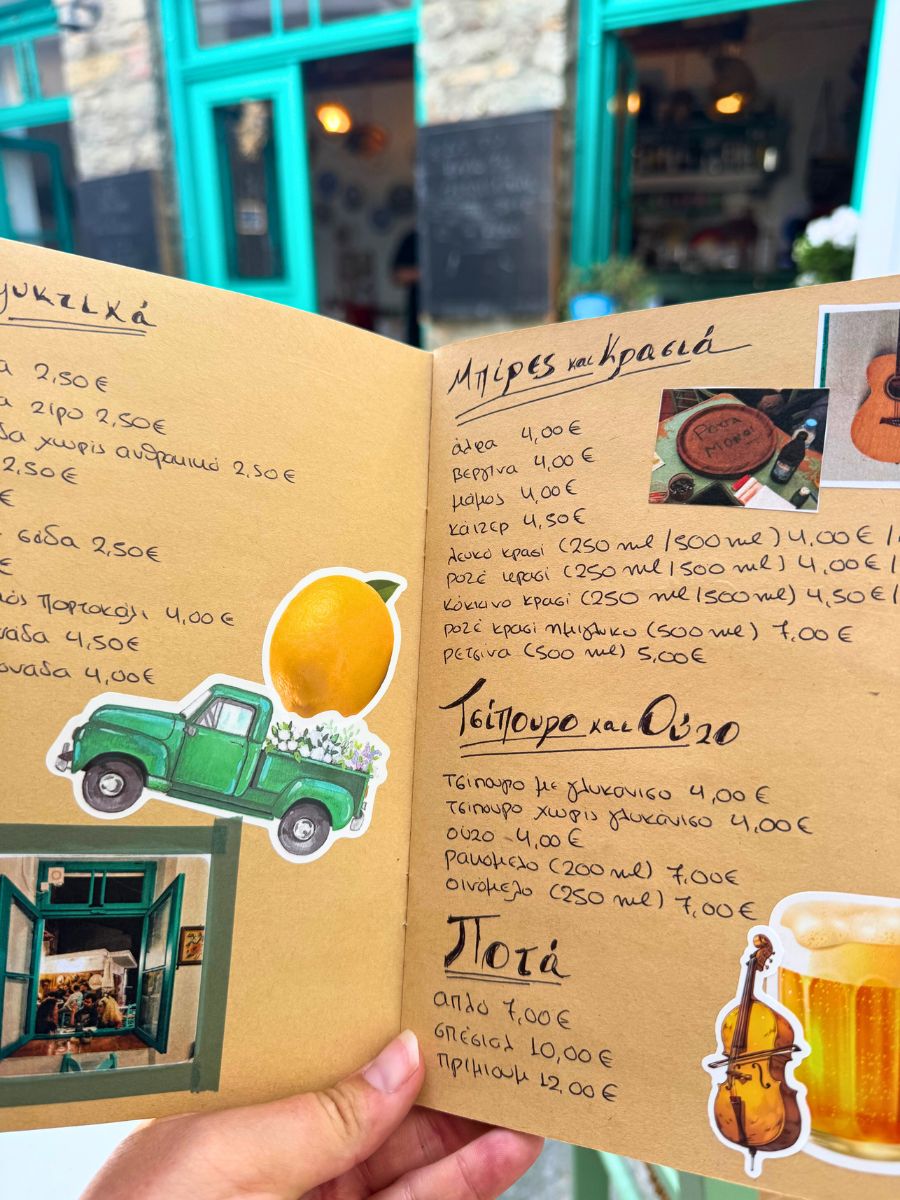Hand holding a Greek café menu with handwritten prices and playful stickers, set against a backdrop of a charming turquoise-door café in Skopelos.