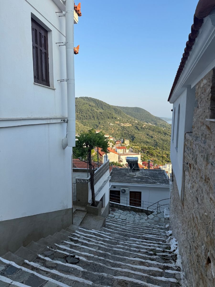Stone staircase between traditional white buildings leading down to a scenic view of green hills and rooftops in Skopelos, Greece.