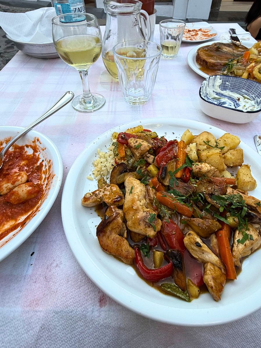 Traditional Greek dinner spread on an outdoor table, featuring roasted chicken with vegetables and potatoes, shrimp in tomato sauce, a jug of white wine, and assorted side dishes.