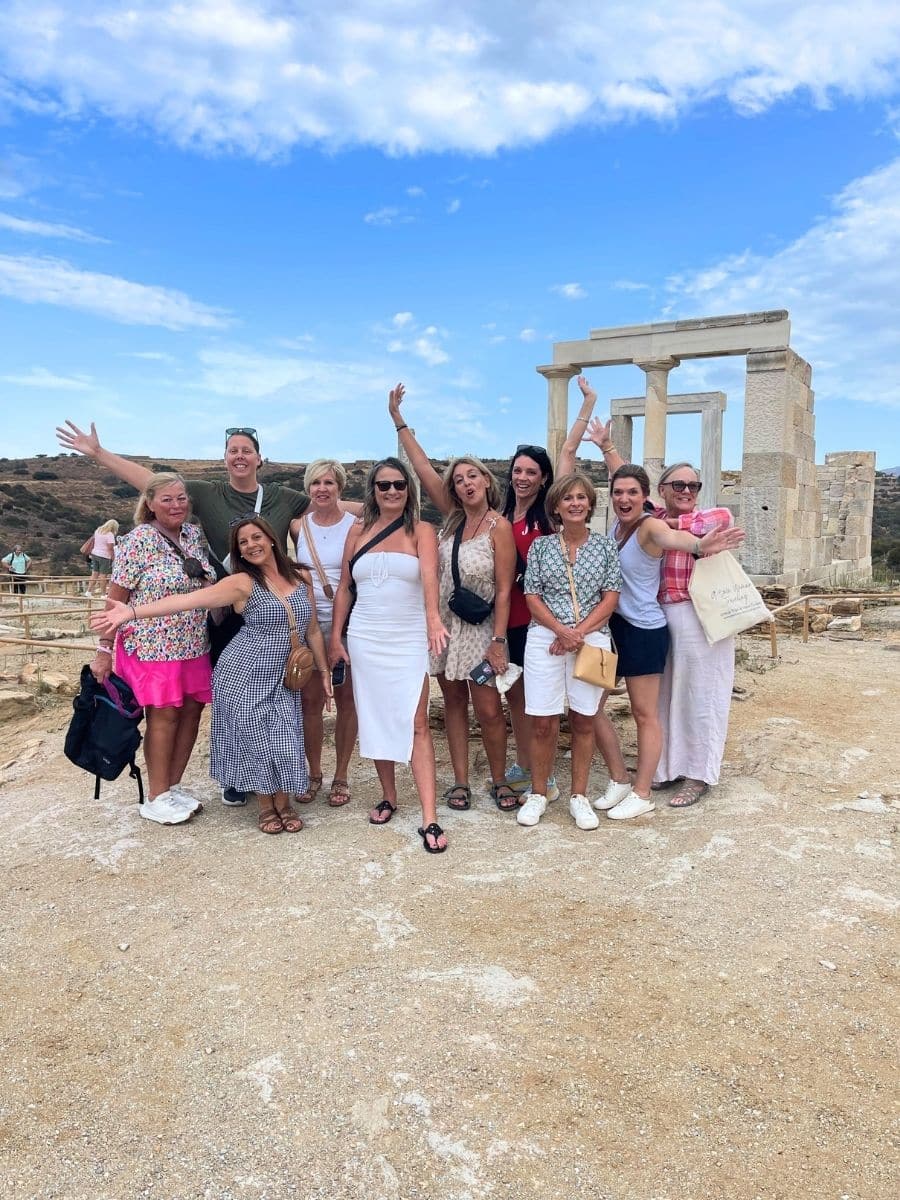 A lively group of travelers poses joyfully in front of ancient Greek ruins under a vibrant blue sky. Their energy and smiles reflect the excitement of exploring historical landmarks together.