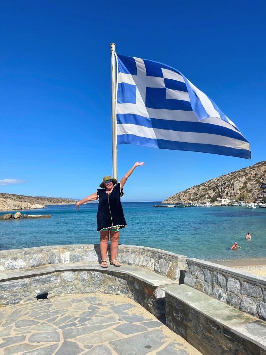 A person standing joyfully next to a large Greek flag on a stone platform overlooking a beautiful seaside. The clear blue water, sandy beach, and rocky hills create a scenic background under a bright blue sky.