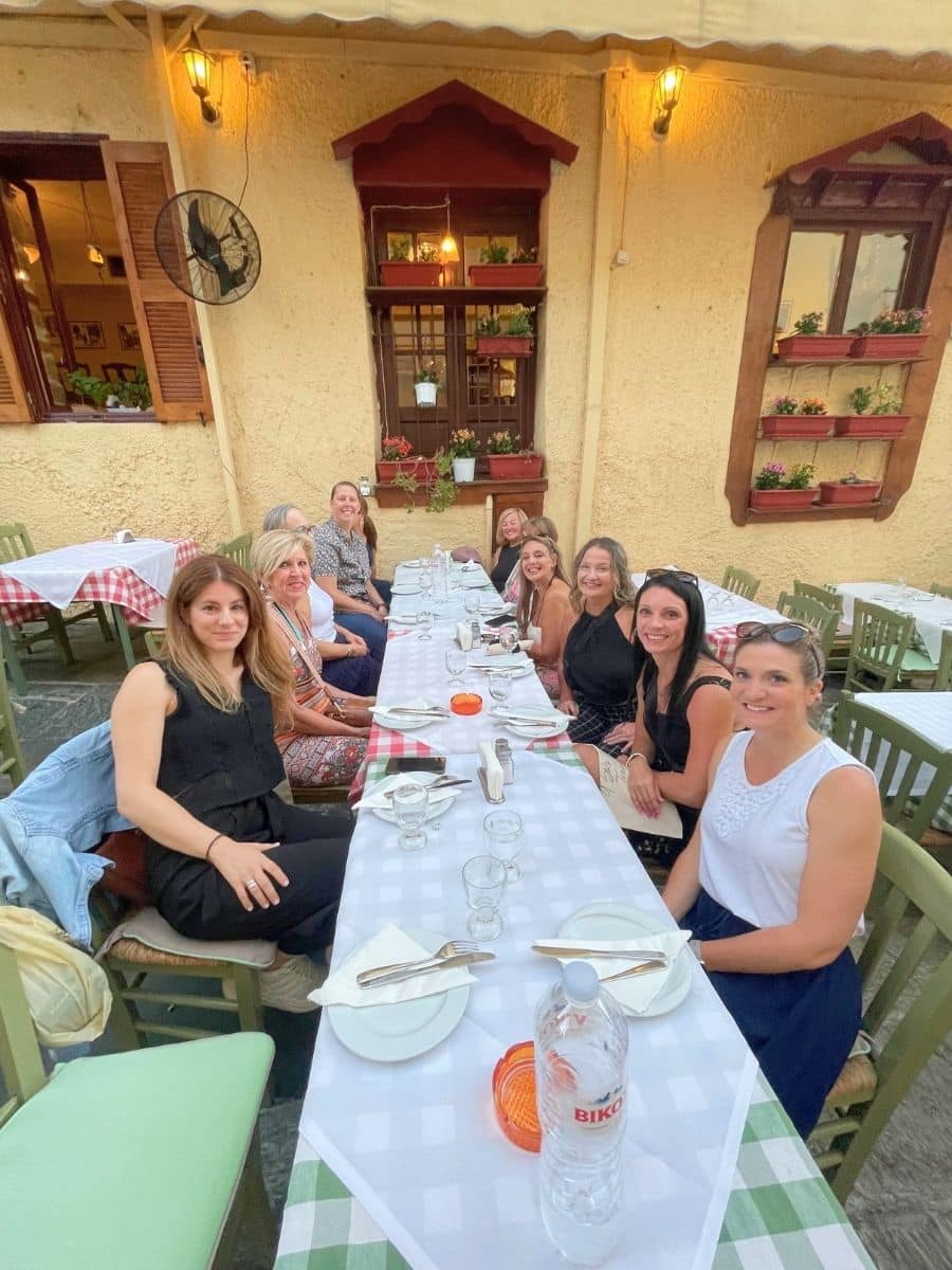 A group of women sitting together at a long outdoor dining table with a Mediterranean-style restaurant backdrop, featuring potted plants and warm lighting.