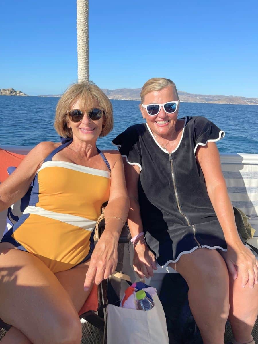 Two women smiling and relaxing on a boat under bright sunlight, with a calm blue sea and distant hills in the background.