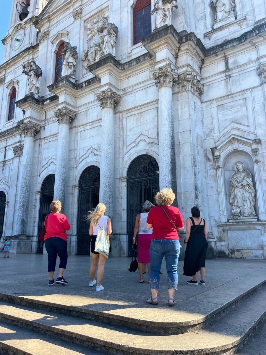 A group of travelers approaches the grand entrance of a historic church, showcasing its ornate architectural details, including towering columns and intricate statues. The steps leading to the church add to its majestic appearance, while the travelers admire the beauty and history of the site.