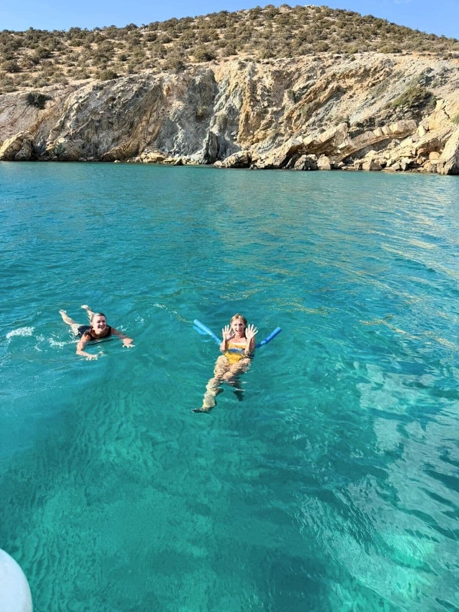 Two people enjoying a refreshing swim in crystal-clear turquoise waters near a rocky coastline. The rugged terrain of the hills in the background contrasts beautifully with the serene, vibrant blue of the sea, creating a tranquil and inviting scene.