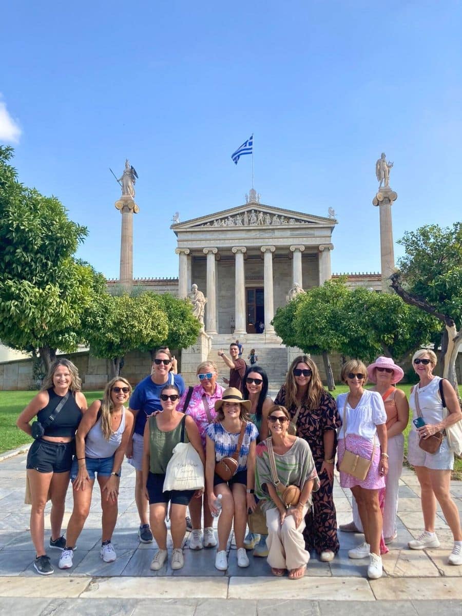 A vibrant group of travelers poses in front of a neoclassical building with Greek columns, adorned with statues and the Greek flag fluttering atop. The bright day enhances the cheerful atmosphere as the group enjoys their cultural adventure.