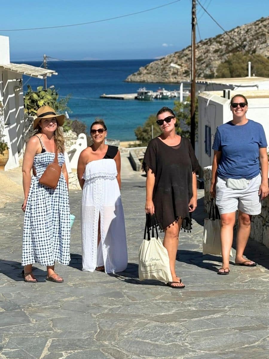 A group of women enjoys a leisurely stroll down a charming cobblestone street with the sparkling Aegean Sea as their backdrop. Their relaxed and stylish summer attire perfectly complements the serene coastal setting of this idyllic Greek island village.