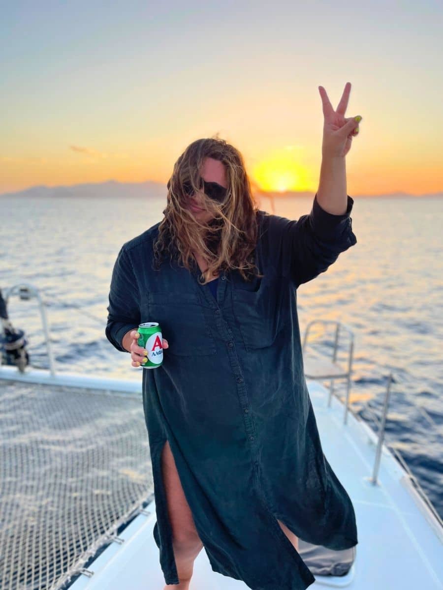A woman in a dark long dress stands on the deck of a boat, holding a can of beer and flashing a peace sign with her other hand. Her hair blows in the wind, partially covering her face, as the sun sets over the sea, creating a golden glow on the water and distant mountains.