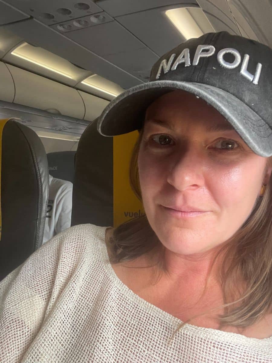 Woman on a plane wearing a black Napoli hat, with a relaxed expression and airplane seating in the background.