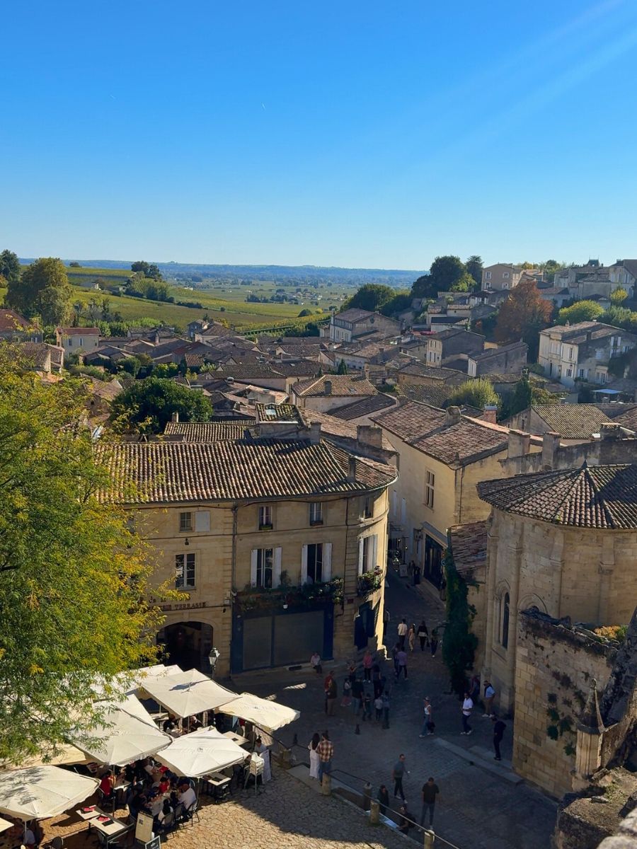 View over the historic village of Saint-Émilion, France, showing stone buildings with terracotta roofs, a sunny café terrace filled with people, and rolling vineyard hills in the distance under a clear blue sky.