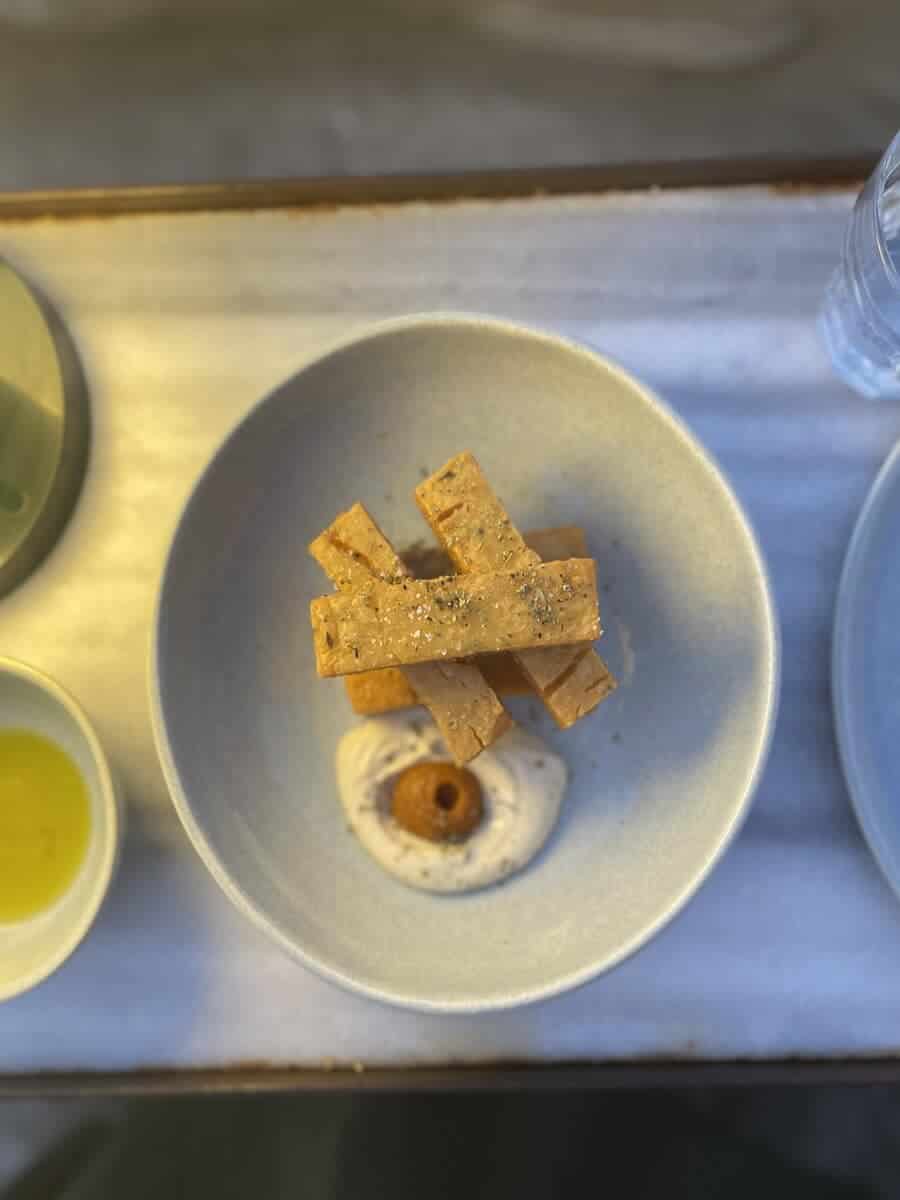 The image shows a minimalist dish served in a light-colored bowl. The dish consists of several rectangular pieces of crisp bread or crackers, arranged neatly on top of a creamy spread or dip. A small round component, possibly an olive or similar garnish, is placed at the center of the spread, giving the dish an elegant and simple presentation. The lighting and composition emphasize the refined, gourmet style of the meal.