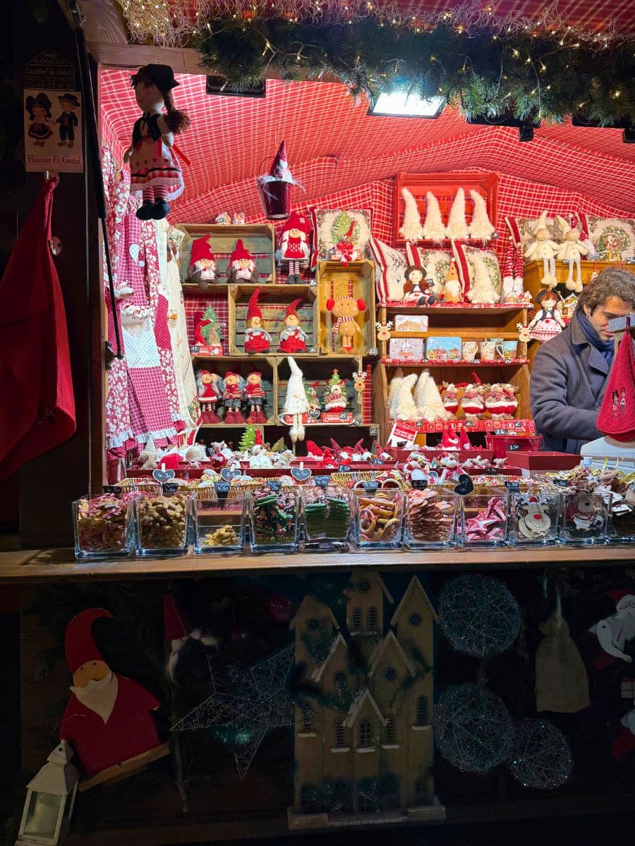A charming market stall at the Strasbourg Christmas Market displaying an assortment of handcrafted holiday decorations, festive figurines, and jars of colorful sweets. The warm red-and-white decor, along with strings of greenery and lights, creates a cozy and cheerful holiday atmosphere. A vendor is seen arranging items.