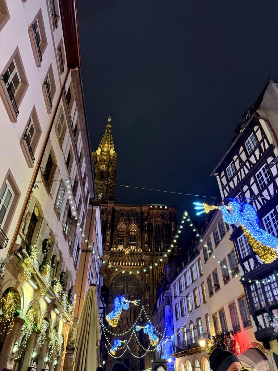 A magical nighttime view of Strasbourg's Christmas market, featuring the majestic Strasbourg Cathedral illuminated in the background. The street is lined with festive decorations, including glowing angel lights and buildings adorned with holiday garlands.