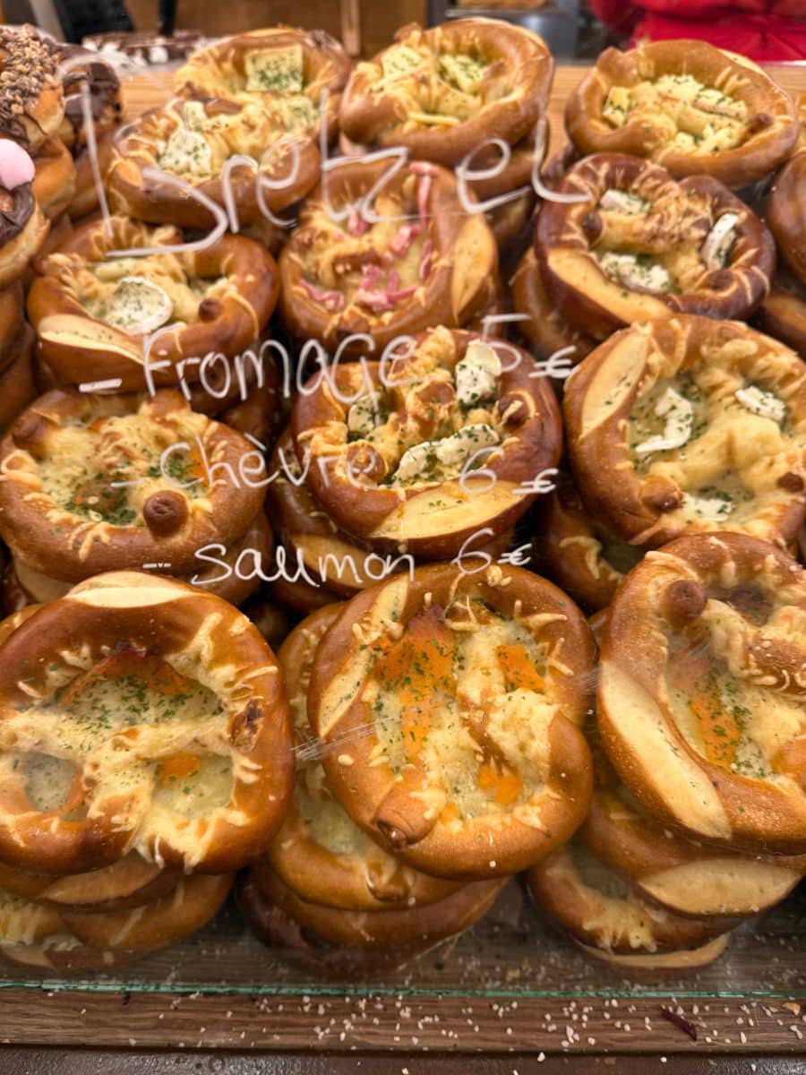A display of freshly baked pretzels with various toppings, labeled with different flavor options: "Fromage" (cheese) for 5€, "Chèvre" (goat cheese) for 6€, and "Saumon" (salmon) for 6€. The pretzels are stacked behind a glass counter at a Christmas market.