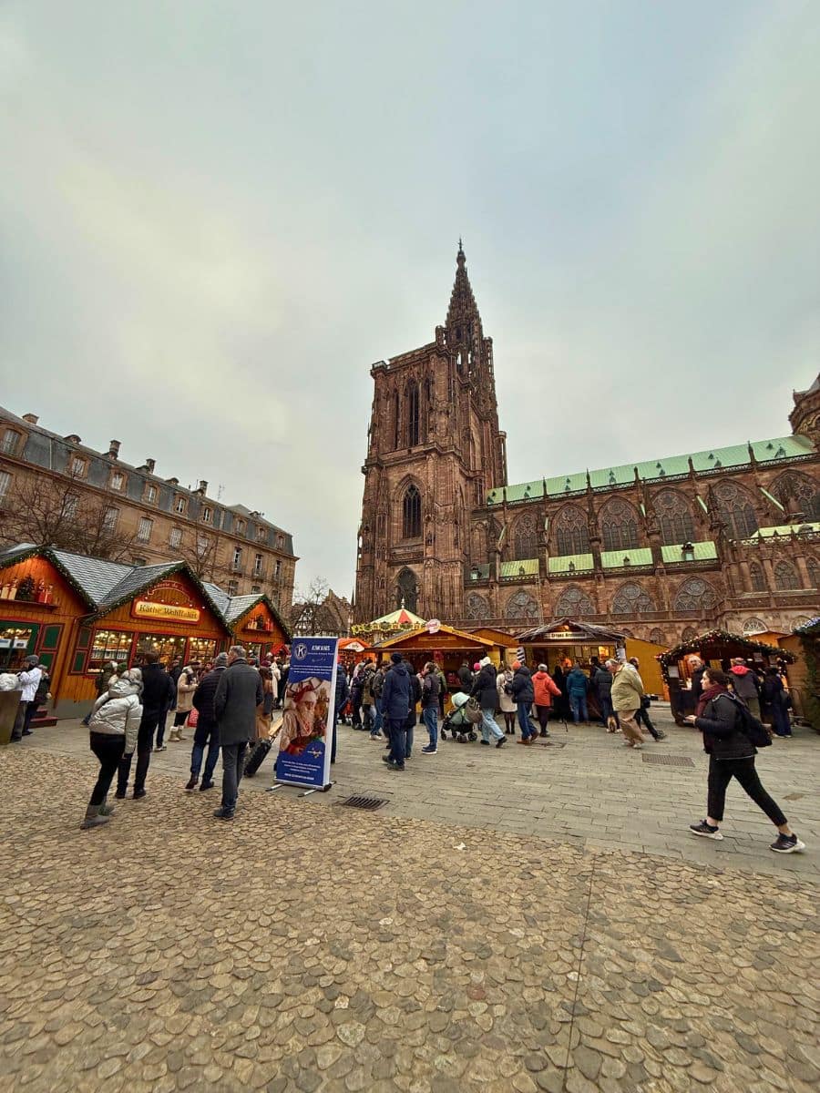 View of the Strasbourg Christmas Market with vibrant holiday stalls and visitors in front of the towering Gothic Strasbourg Cathedral, creating a festive and historic atmosphere.
