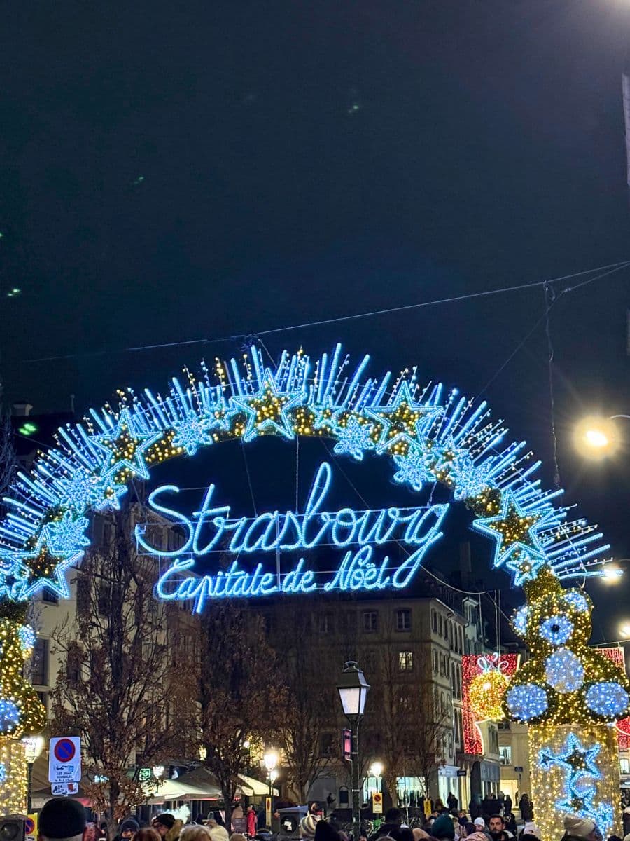 A brightly lit archway decorated with glowing blue and gold stars and ornaments, featuring the text "Strasbourg Capitale de Noël" in elegant script. The festive display is surrounded by trees adorned with lights, and a lively crowd of people is gathered beneath the arch. The background showcases buildings illuminated by warm streetlights on a festive evening.