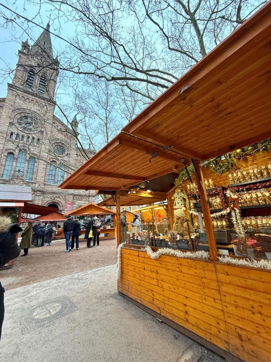 Wooden market stalls adorned with festive decorations at a Christmas market in Strasbourg. The background features a grand historic church with a tall spire, and bare tree branches frame the scene under a light blue sky. People stroll through the market, browsing the offerings.