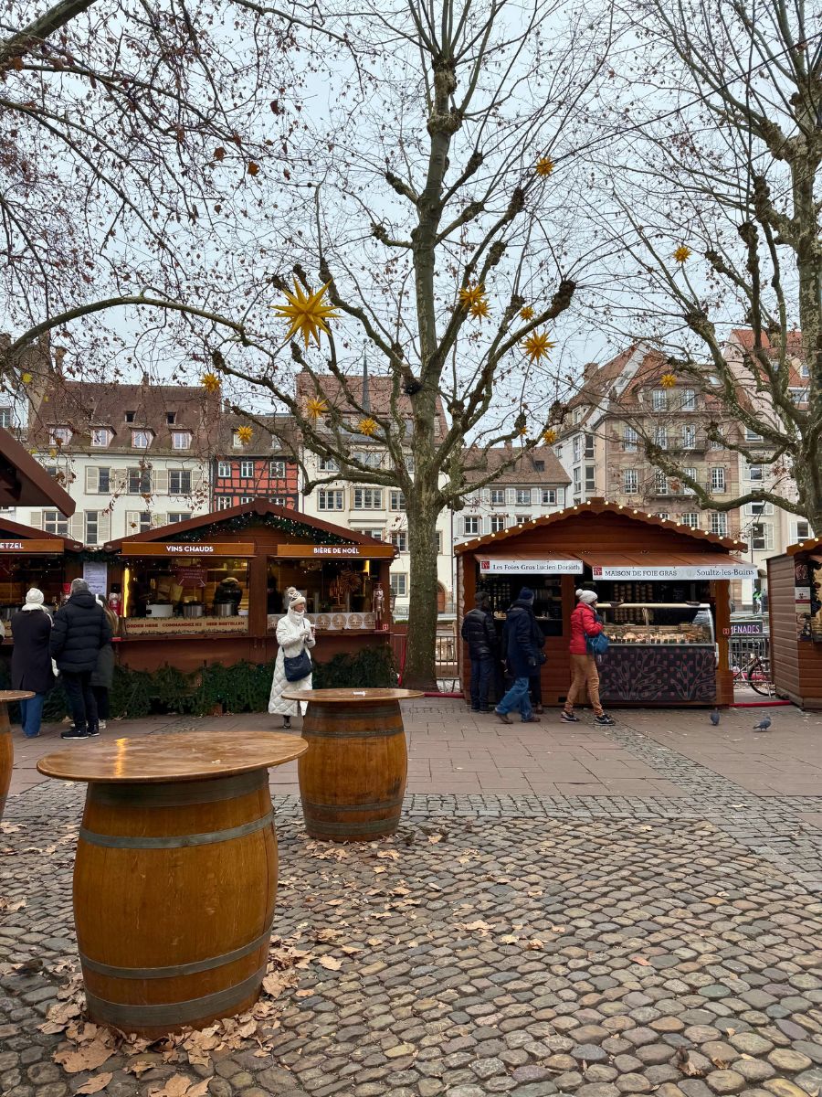 A peaceful corner of the Strasbourg Christmas market with wooden barrel tables on cobblestone streets, surrounded by charming wooden stalls offering festive treats like mulled wine and Christmas beer. Golden star decorations hang from bare trees, adding a festive sparkle.
