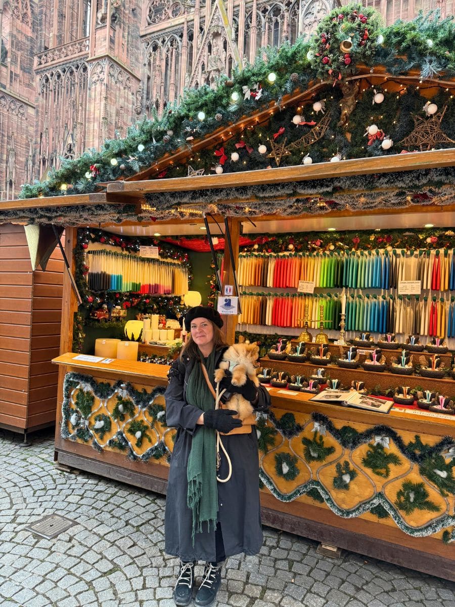 A festive market stall in Strasbourg decorated with evergreen garlands, ornaments, and string lights. The stall displays colorful candles and small handcrafted items. In front of it, a person wearing a long coat, scarf, and beret poses with a fluffy dog. The cobblestone street and the intricate façade of Strasbourg Cathedral in the background add to the charming holiday atmosphere.
