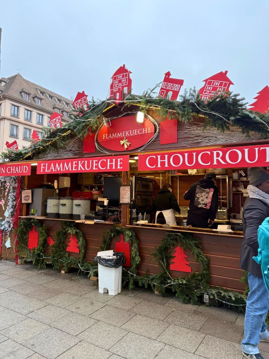 A charming food stall at the Strasbourg Christmas Market, specializing in Alsatian delicacies like Flammekueche (a thin, pizza-like dish topped with crème fraîche, onions, and lardons) and choucroute (a traditional sauerkraut dish). The festive decorations, including evergreen garlands and red cutouts of houses and Christmas trees, add a warm holiday vibe. Did you try anything from here? Flammekueche is a must-have if you’re exploring Alsace!