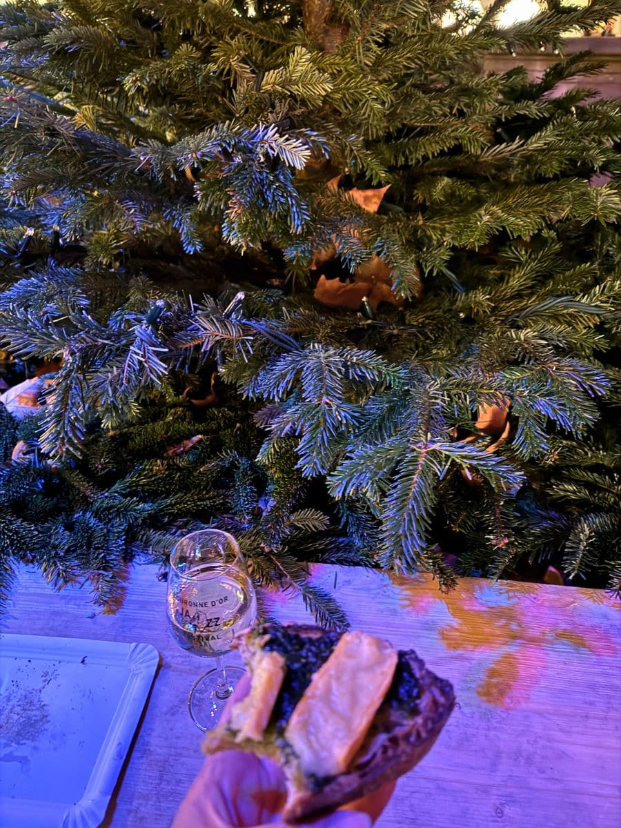 A festive scene featuring a decorated Christmas tree, a glass of white wine, and a slice of toasted bread topped with cheese and greens. The warm, colorful lighting adds to the holiday atmosphere.