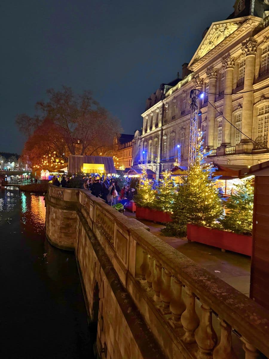 A lively Christmas market along the water, with festive lights illuminating the historic building and decorated trees. Reflections of the lights shimmer on the canal, creating a warm and enchanting evening scene.