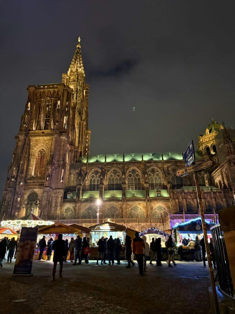 A nighttime view of Strasbourg Cathedral illuminated against the dark sky, showcasing its intricate Gothic architecture. At the base, a bustling Christmas market is set up with wooden stalls decorated with lights, offering festive goods and treats. Visitors are seen enjoying the vibrant atmosphere.