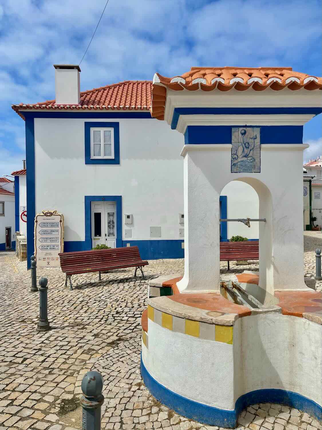 A charming square in Ericeira, Portugal, featuring a traditional whitewashed building with blue trim and a red-tiled roof. In the foreground, a small fountain with an arched structure is highlighted, surrounded by cobblestone pavement. A wooden bench and a sign are visible near the building, all set against a partly cloudy sky.
