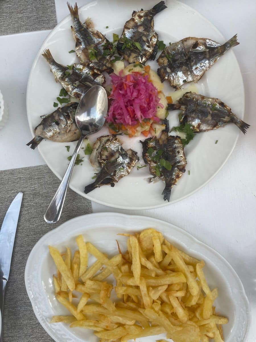 A delicious Mediterranean meal served on a white table. The main dish features grilled sardines arranged in a circular pattern on a white plate, garnished with fresh herbs and accompanied by a side of potato salad topped with vibrant pickled red cabbage. A spoon rests on the plate. Alongside, a separate white dish holds a portion of golden, crispy French fries. The table setting includes a knife and a textured gray placemat, enhancing the rustic yet elegant dining experience.