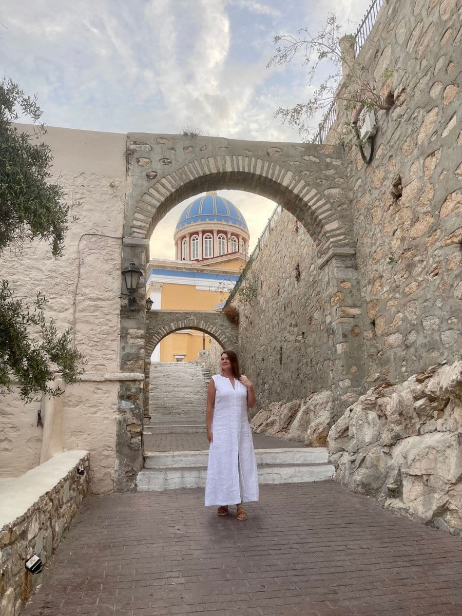 Melissa in a white linen dress stands on a stone-paved pathway, surrounded by historic stone walls and archways. Behind her, a grand church with a blue dome and yellow accents rises above the scene, illuminated by the soft evening light. The pathway leads up a series of white stone steps, framed by rustic walls adorned with lanterns and creeping vines. The atmosphere is peaceful and timeless, capturing the charm of Syros, Greece.
