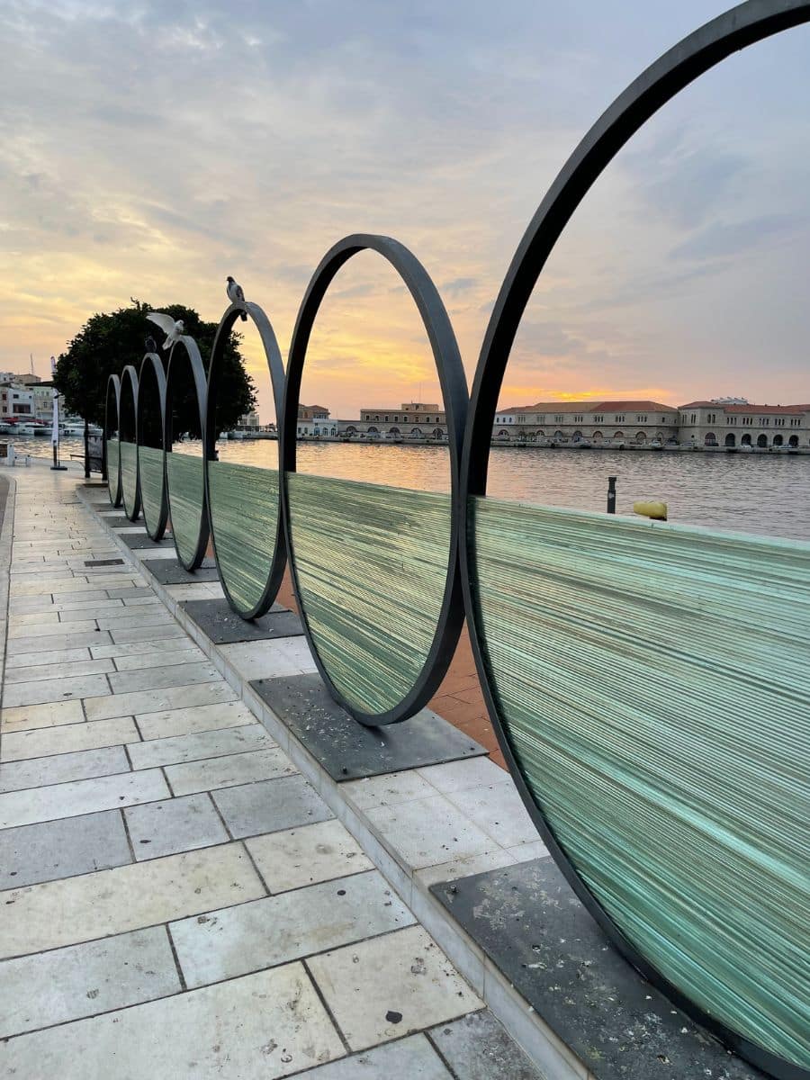 A modern waterfront art installation in Syros, Greece, featuring large metal rings with glass panels strung with delicate horizontal lines, creating a translucent barrier along the promenade. Two pigeons perch on the rings as the golden hues of the sunset cast a warm glow over the harbor. In the background, historic buildings with arched windows line the waterfront, reflecting the island's rich maritime heritage. The paved walkway, adorned with natural stone tiles, leads towards the serene waters, blending contemporary design with the timeless beauty of the Cycladic landscape.