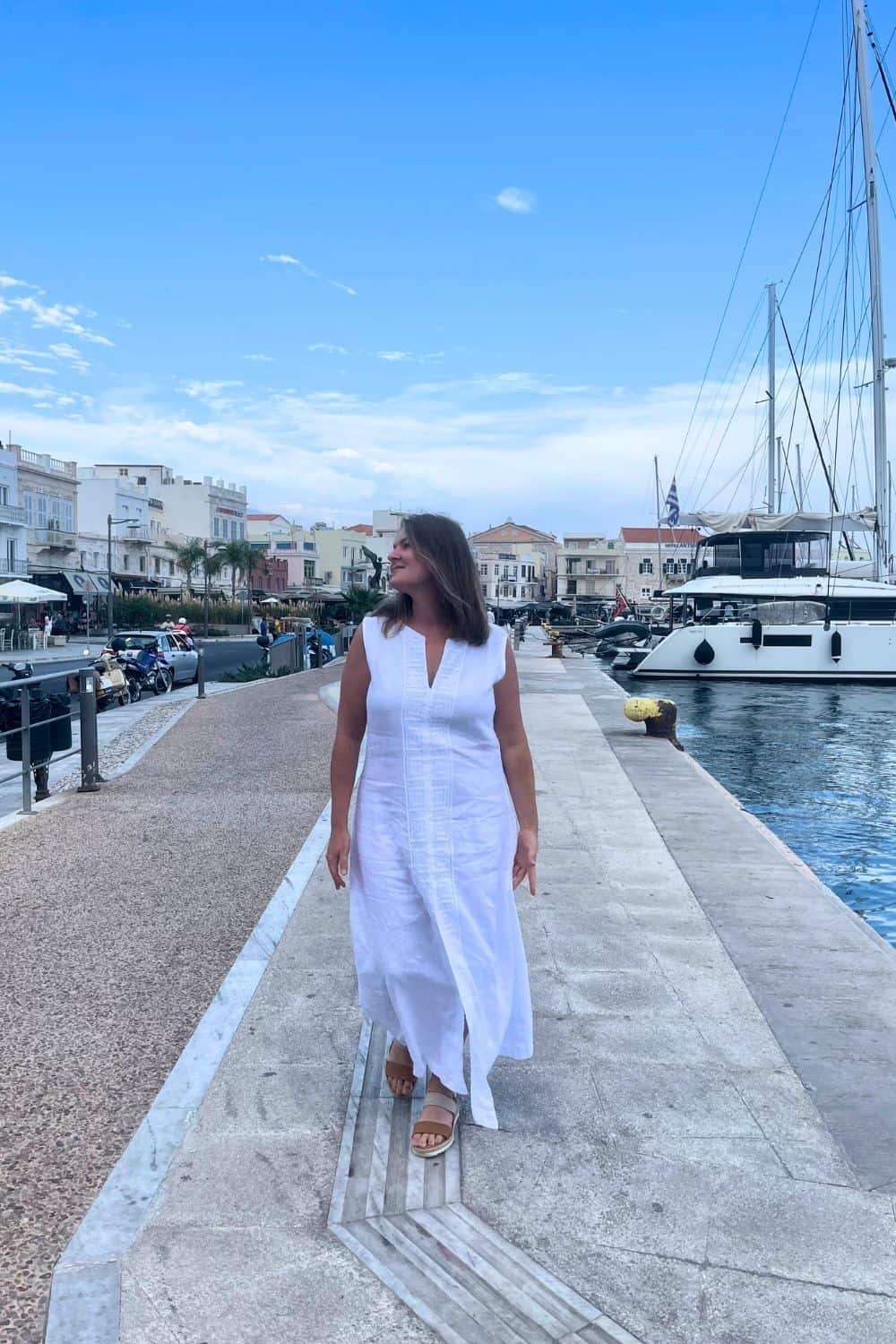 A solo female traveler in a white dress walks along a marina on one of the hottest Greek islands in May, with sailboats docked and the town's architecture visible in the background.