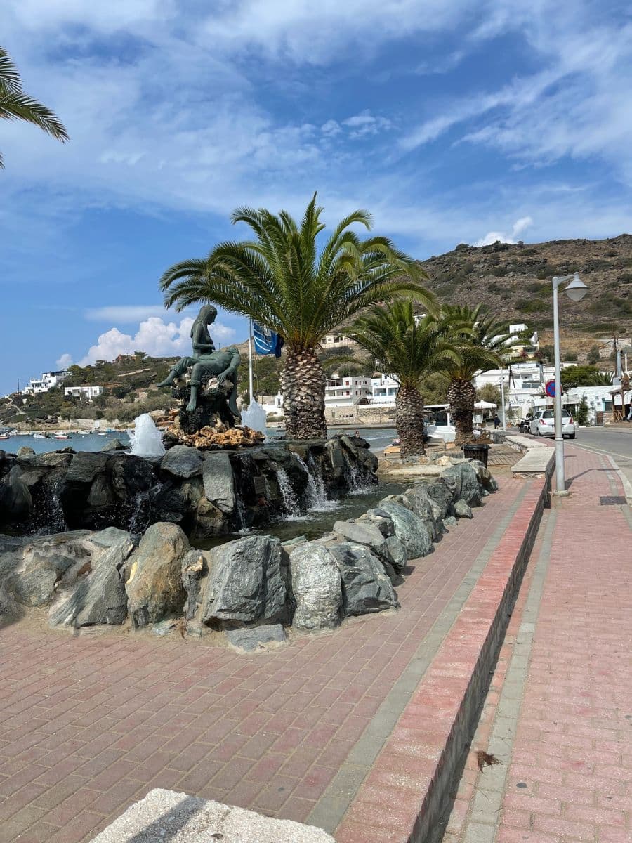 A picturesque seaside promenade on the island of Syros, Greece, featuring a mermaid fountain with cascading water set against a backdrop of palm trees, white houses, and rolling hills. The blue sky with scattered clouds enhances the coastal charm, while the red-brick pedestrian path curves alongside the waterfront, inviting leisurely strolls.