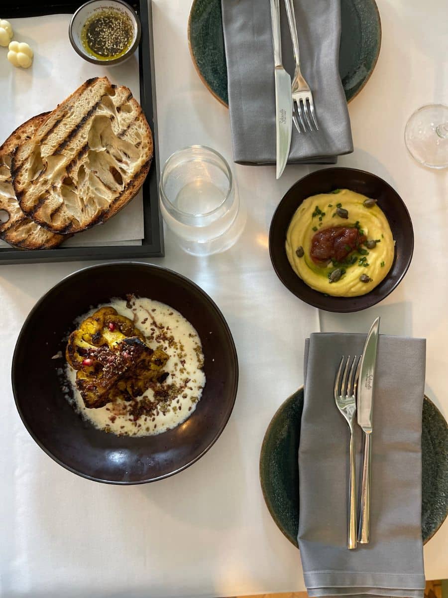 A beautifully plated meal at a fine dining restaurant, featuring grilled sourdough bread with olive oil, a dish of creamy mashed fava beans topped with capers and caramelized onion, and a roasted cauliflower steak served with a velvety sauce and garnished with pomegranate seeds. The table setting includes elegant cutlery, gray napkins, and dark ceramic plates, all arranged on a crisp white tablecloth.