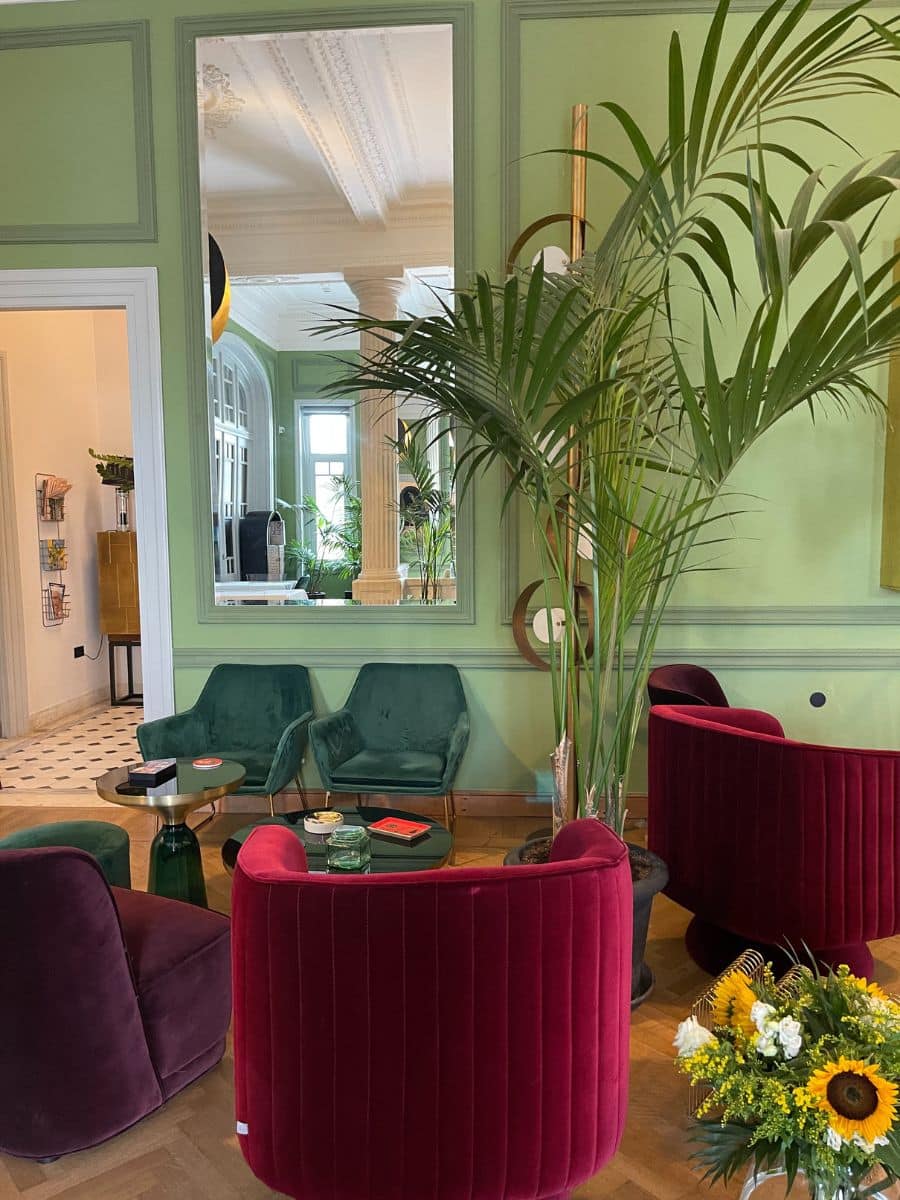 A stylish interior lounge with green paneled walls, large mirrors, and a high ceiling with intricate molding. The room is furnished with plush emerald green and deep burgundy velvet chairs arranged around gold and green coffee tables. A tall potted palm plant adds a tropical touch, while a bouquet of sunflowers and white flowers sits in the foreground. The space has a sophisticated yet cozy ambiance, blending classic architecture with modern decor.