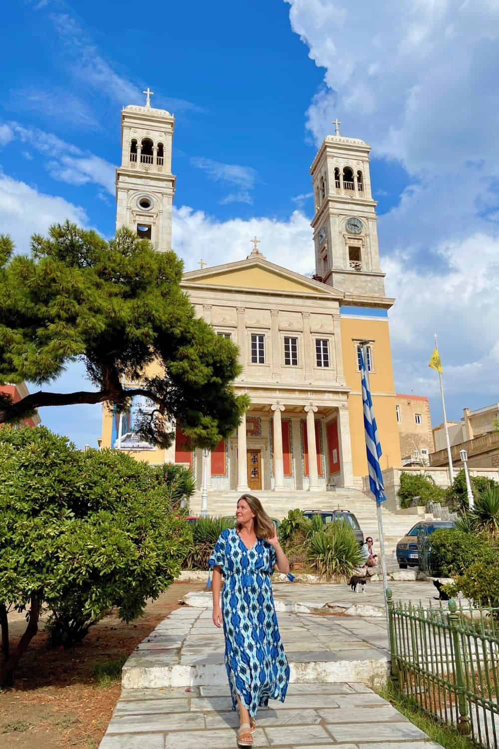 A woman in a flowing blue and white patterned dress walks confidently in front of the striking yellow and white facade of a neoclassical church with twin bell towers under a bright blue sky. Lush greenery and the vibrant colors of the national flag of Greece complement the historic architecture, creating a picturesque scene on the island of Syros.
