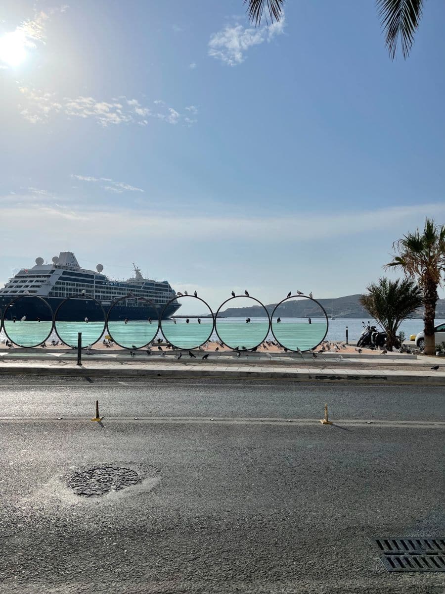 A scenic view of a coastal area with a large cruise ship docked near the shore. In the foreground, a series of circular metal frames line the waterfront promenade, with pigeons perched on them. The calm turquoise sea reflects the sunlight, and palm trees add to the tropical ambiance. In the background, rolling hills rise from the water, completing the tranquil maritime scene.