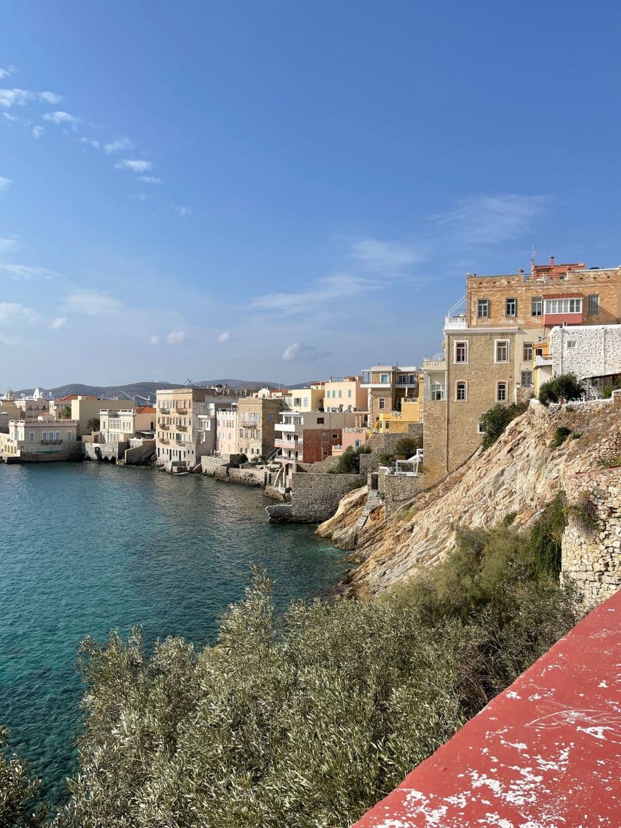 A picturesque coastal scene of Syros, Greece, featuring colorful neoclassical buildings perched on rocky cliffs overlooking the deep blue Aegean Sea. The buildings, painted in warm pastel hues, contrast with the rugged terrain and the clear sky. The calm water gently laps against the stone walls and staircases leading into the sea, creating a serene Mediterranean atmosphere.