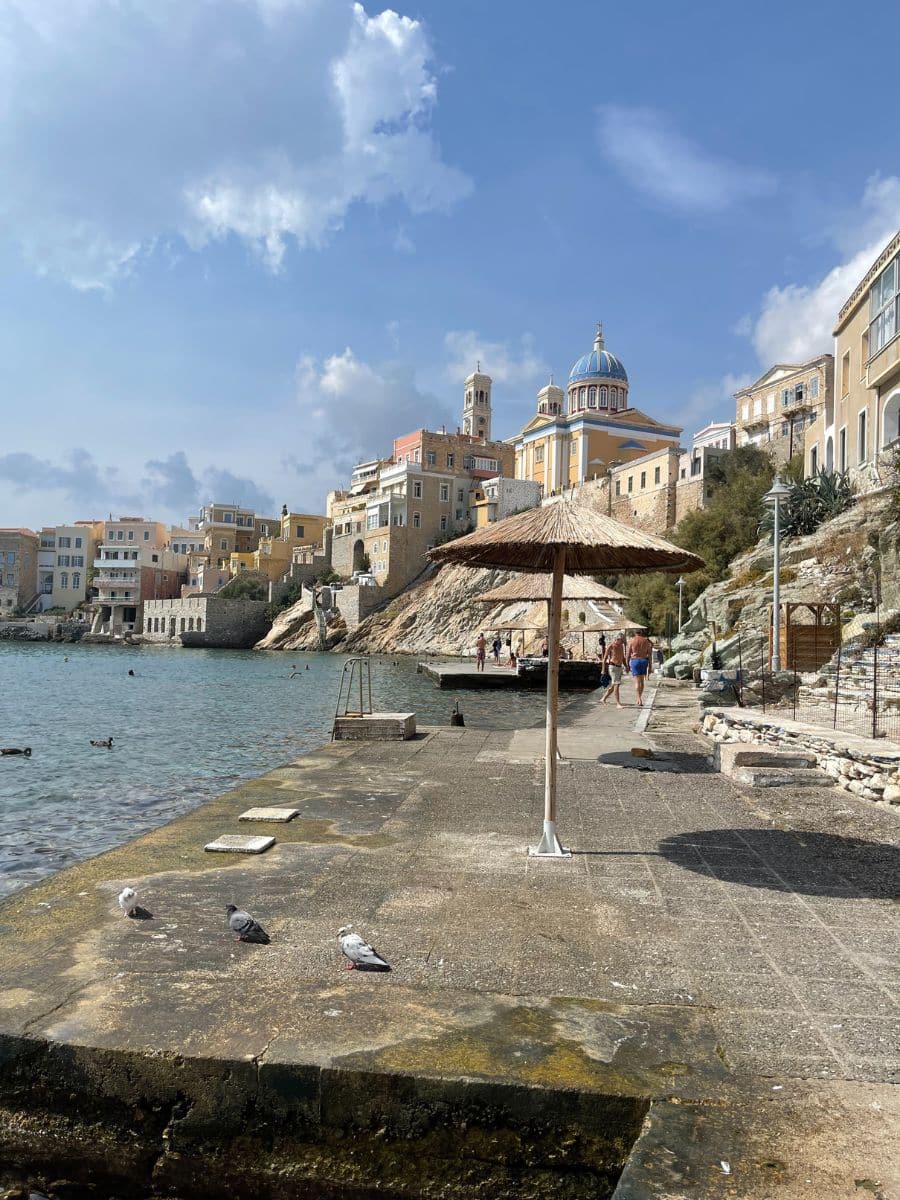A scenic coastal view of a Mediterranean town with pastel-colored buildings perched on rocky cliffs. A blue-domed church with neoclassical architecture stands prominently among the structures. In the foreground, a stone pier extends into the sea, with a thatched umbrella providing shade. Pigeons are pecking at the ground, and people are strolling along the pier, some dressed in swimwear. The calm waters are dotted with a few swimmers. Above, the sky is a mix of blue and white, with large, billowy clouds casting soft shadows over the town.