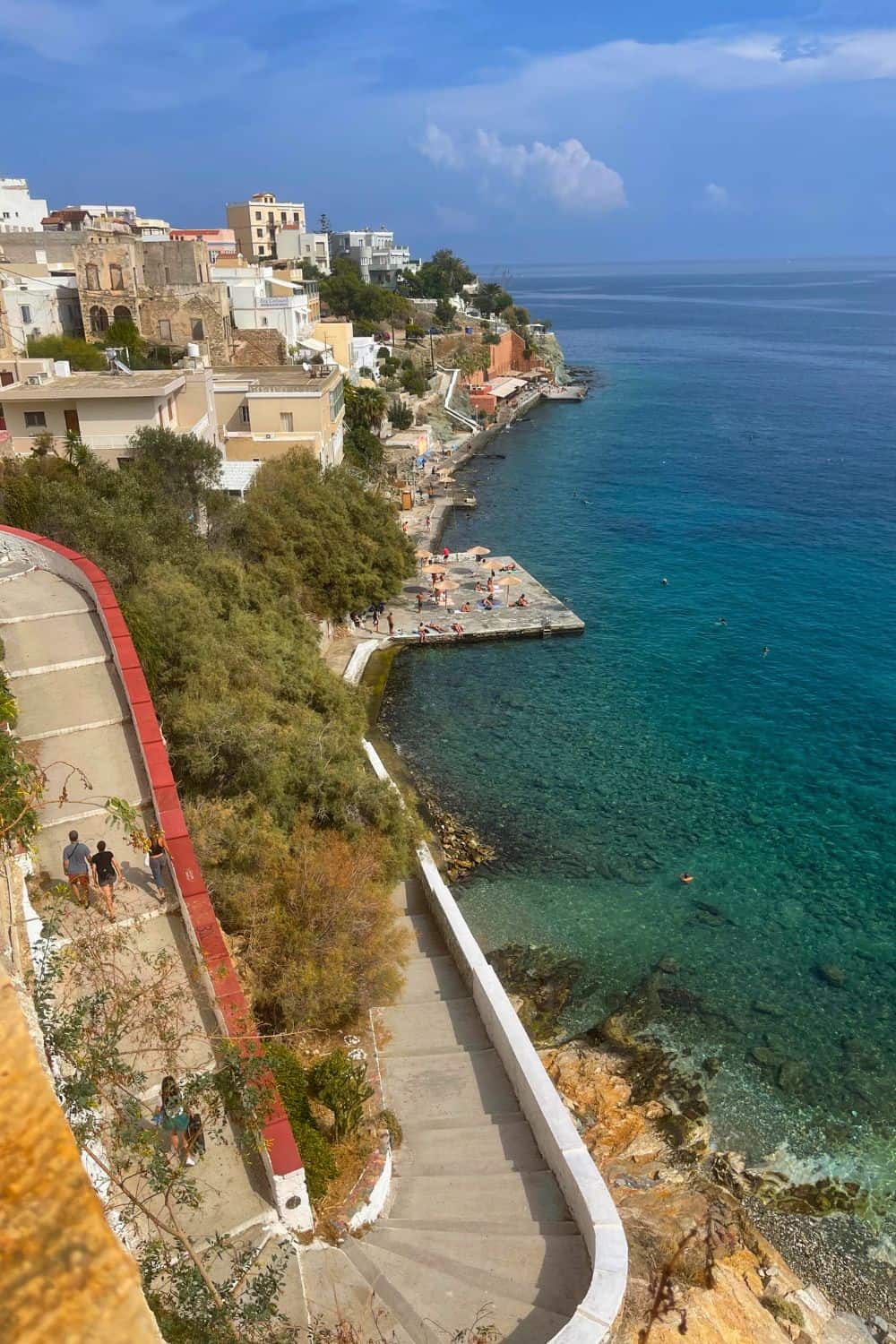  An aerial view of a island of Syros and the seaside promenade on a Greek island, one of the hottest destinations to visit in May, featuring clear blue waters, a meandering coastal path, and traditional Mediterranean buildings.