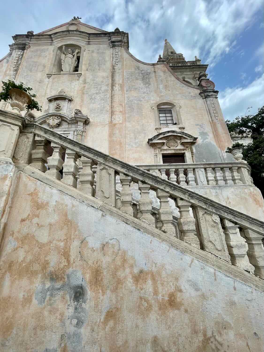 A historic building in Taormina, Sicily, with weathered plaster walls and an ornate staircase leading up to it. The façade includes detailed architectural elements and a statue in a niche, set against a partly cloudy sky.