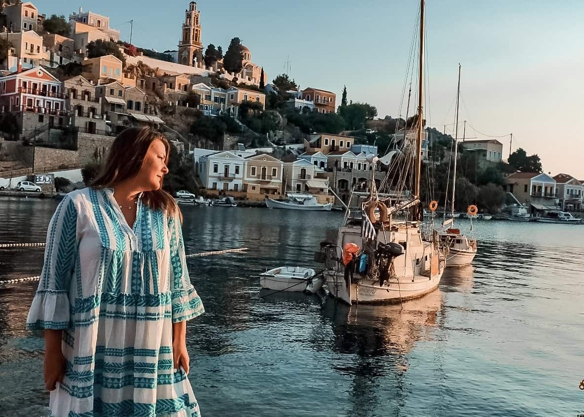 A serene sunset paints the harbor of a picturesque village, where a solo woman traveler in a flowy white and teal dress gazes out over the calm waters, reflecting on her solo journey amidst the backdrop of anchored sailboats and historic hillside architecture.