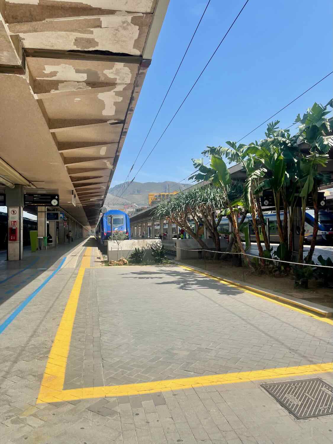 Palermo train station platform in Sicily, with a modern blue train parked at the station. The platform is lined with yellow and blue markings, and lush greenery is visible in the background under a bright blue sky.