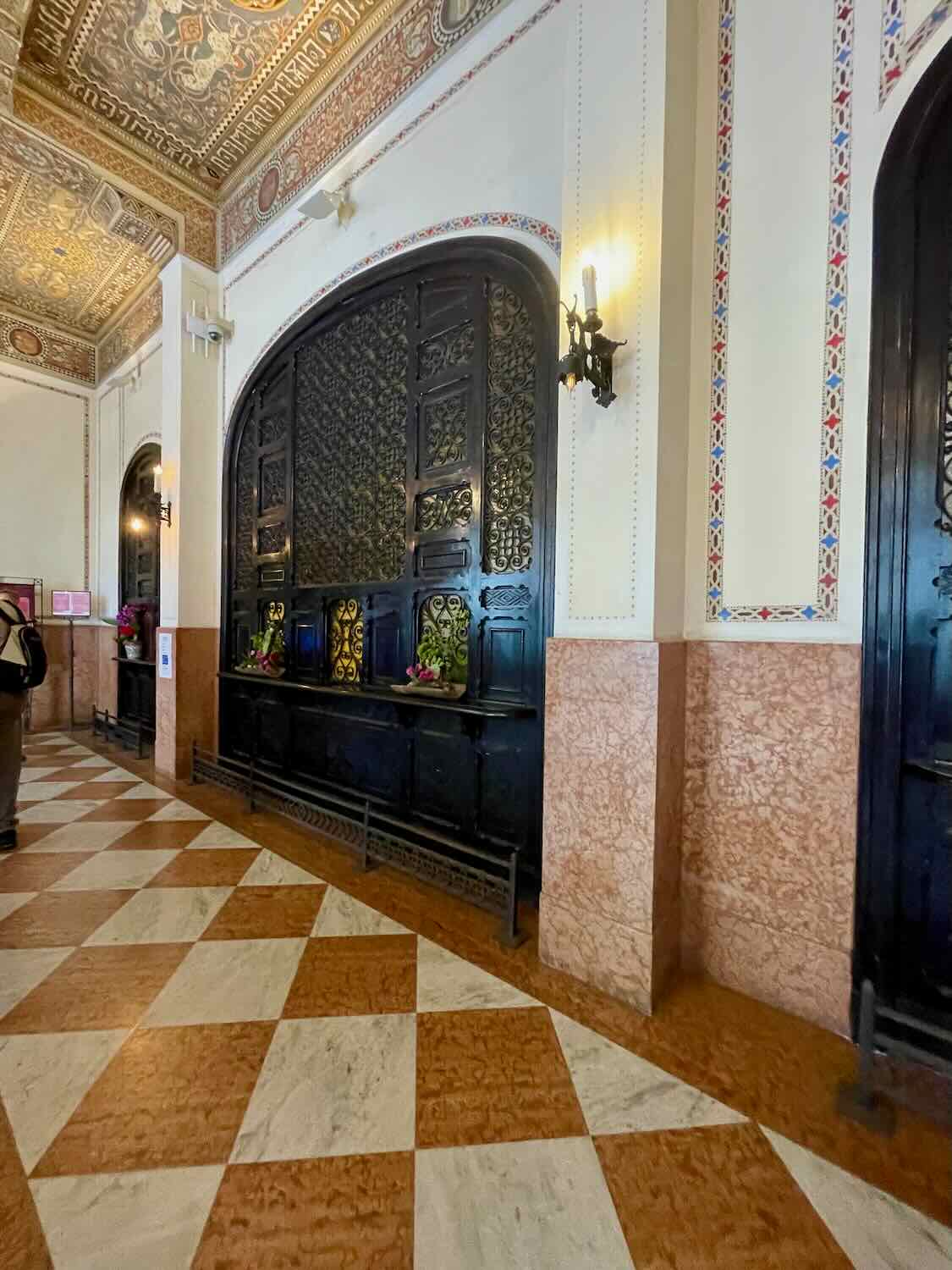 Interior of a train station featuring a grand, ornate black metal fireplace with decorative ceramic tiles. The walls are adorned with intricate tiling and the floor showcases a classic checkered pattern, enhancing the historical ambiance of the space.