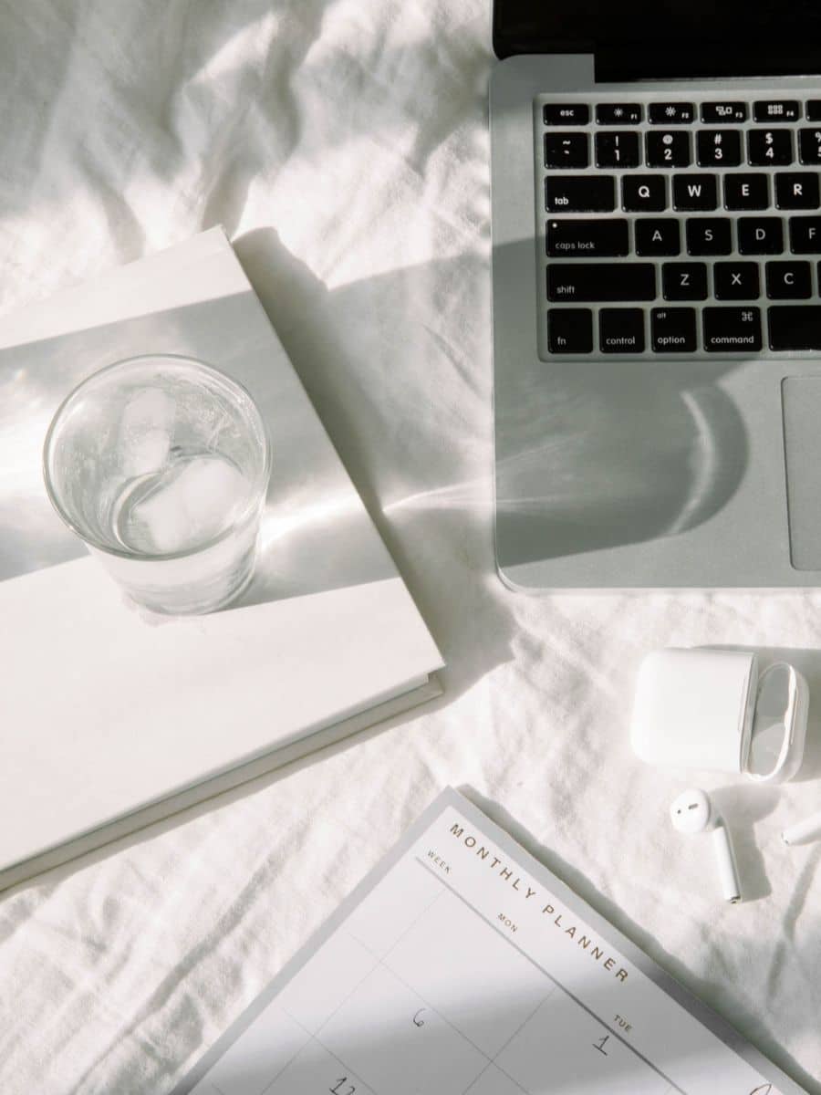Flatlay of laptop, planner, and AirPods on a white bed—showcasing an organized and minimalist setup for travel planning in Notion.